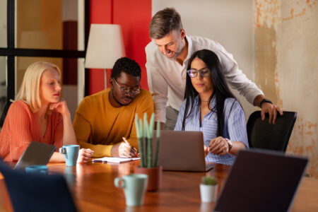 A diverse team collaborates at a wooden table, focused on a laptop and notes, with coffee cups and plants visible in the workspace.