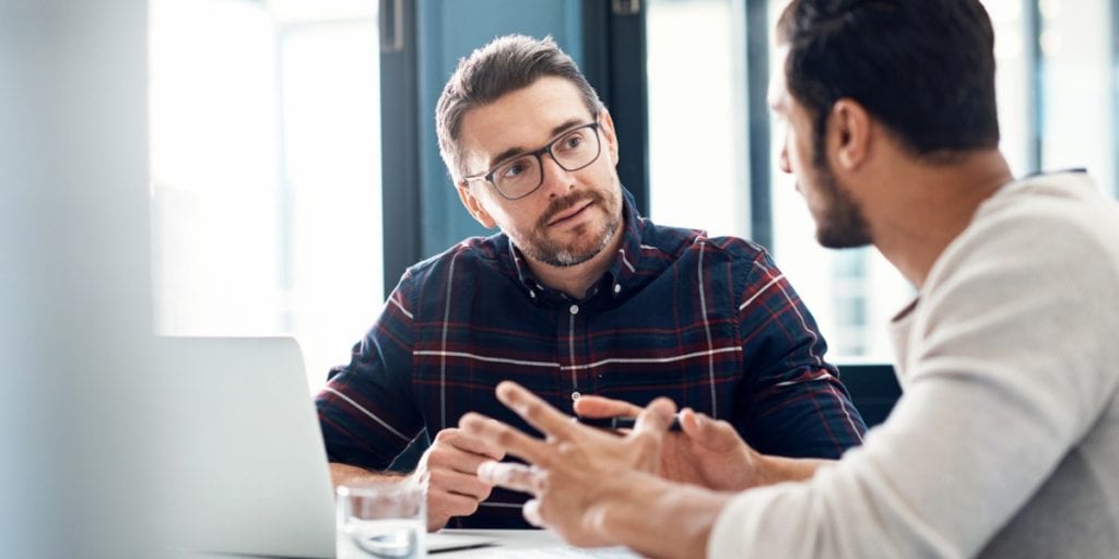 Two men dressed in a smart casual way are conversing in a meeting. They are sat at a desk with a laptop open and pen in hand.