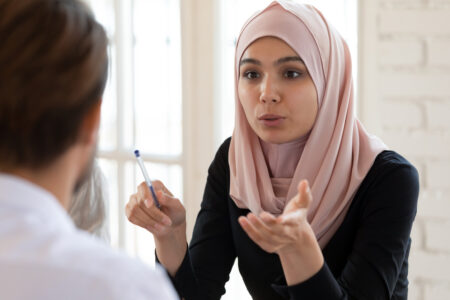 Concentrated serious young arabian businesswoman in hijab talking to male team leader, headshot. Focused mixed race partners discussing marketing strategy or project details together at office.
