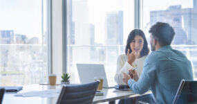 Business woman and man meeting and talking. Both are casually dressed. There is a laptop computer on the table. They are sitting at a boardroom table with a window behind them