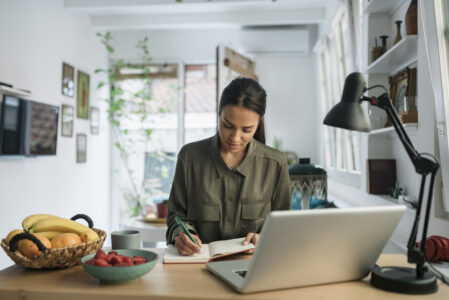 Female employee working from home writing notes on a pad behind a laptop