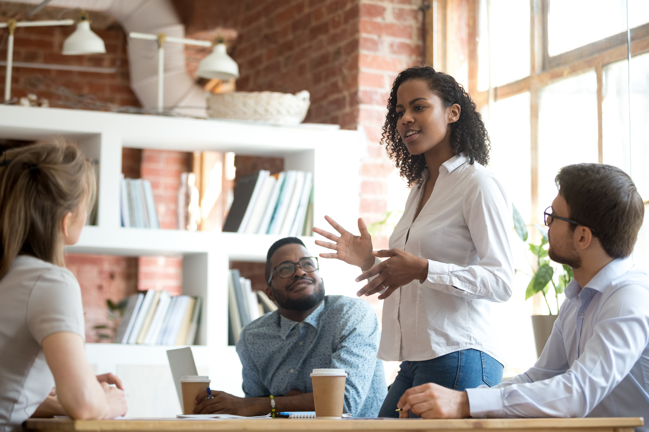 A woman stands at a table, engaged in a discussion with three people, in a bright, modern office with brick walls and bookshelves.