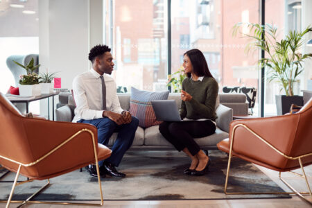A woman in a sweater and a man in a suit sit on a sofa in a modern office, discussing while using a laptop. Stylish decor surrounds them.