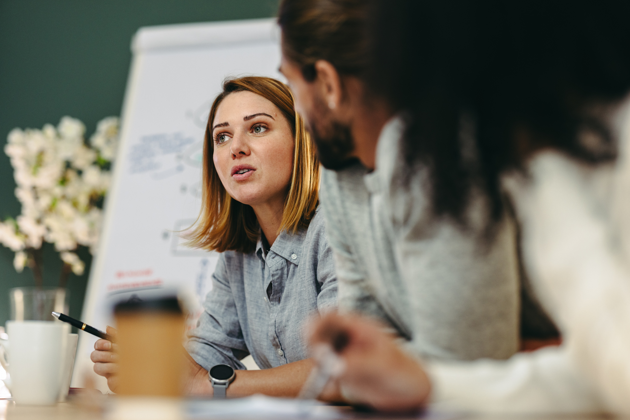 A group of people engaged in a discussion around a table, with a whiteboard and coffee cups in a professional setting.