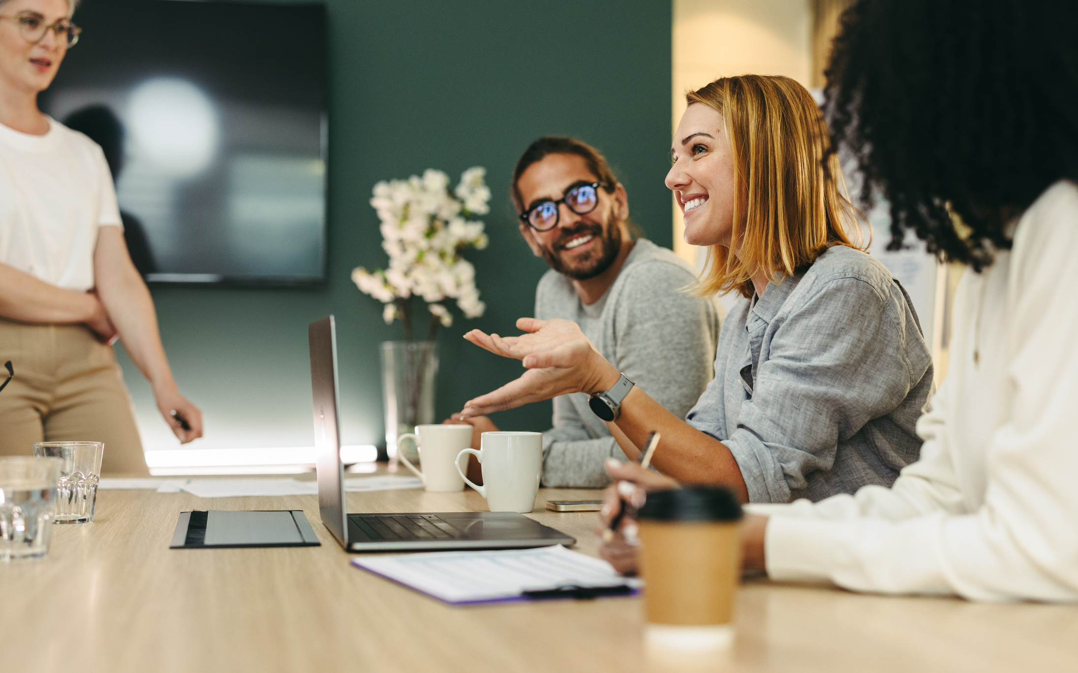 A diverse group of professionals engaged in a meeting at a conference table, with a laptop and coffee cups displayed.