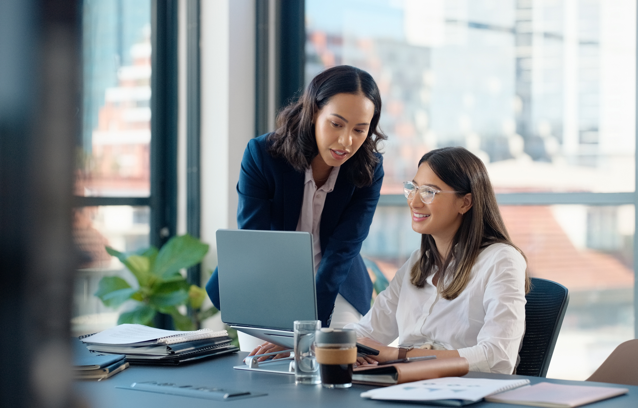 Two women share ideas in a bright office, one pointing at a laptop screen, with plants and notebooks on the desk.