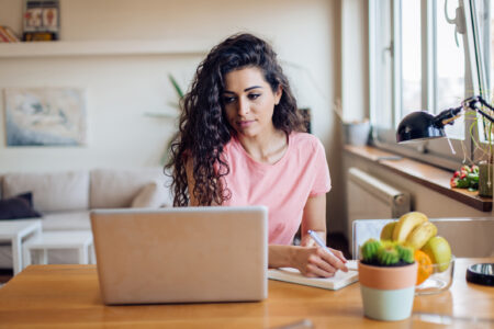 A person with curly hair sits at a desk, writing in a notebook, with a laptop and a bowl of fruit nearby, in a bright room.