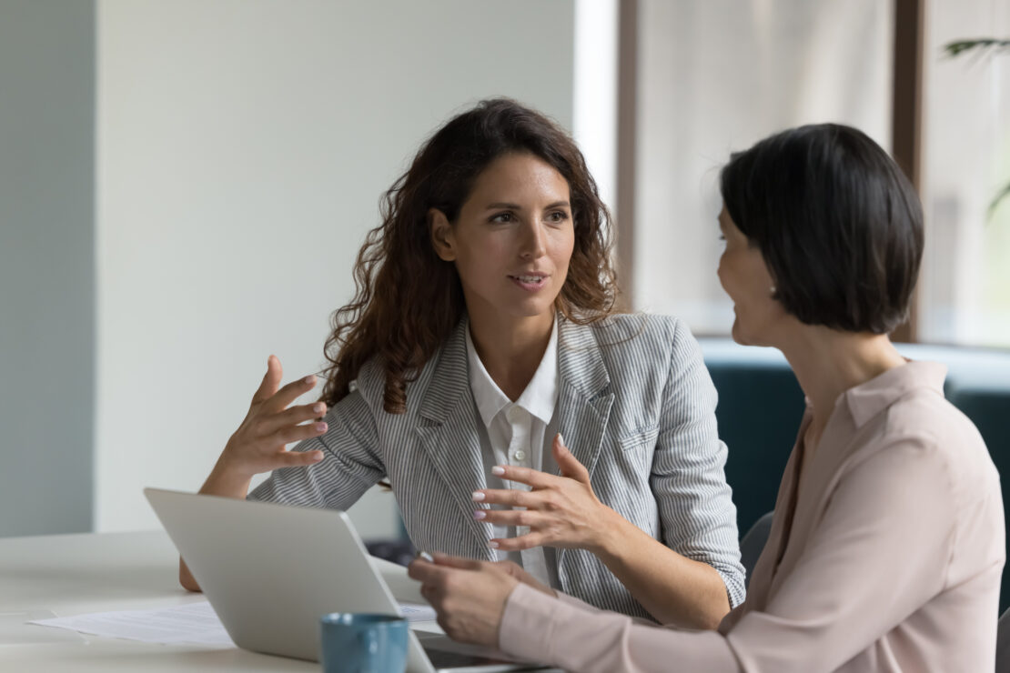 Two women engage in a conversation at a desk with a laptop and a blue coffee mug, surrounded by natural light in a modern office.