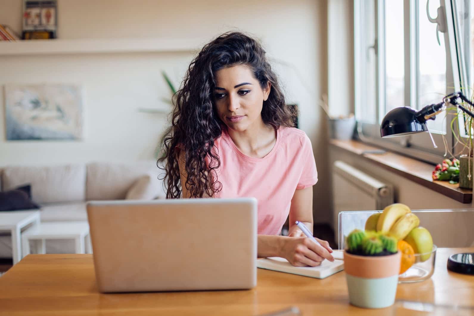 A person with curly hair sits at a desk, writing in a notebook, with a laptop and a bowl of fruit nearby, in a bright room.