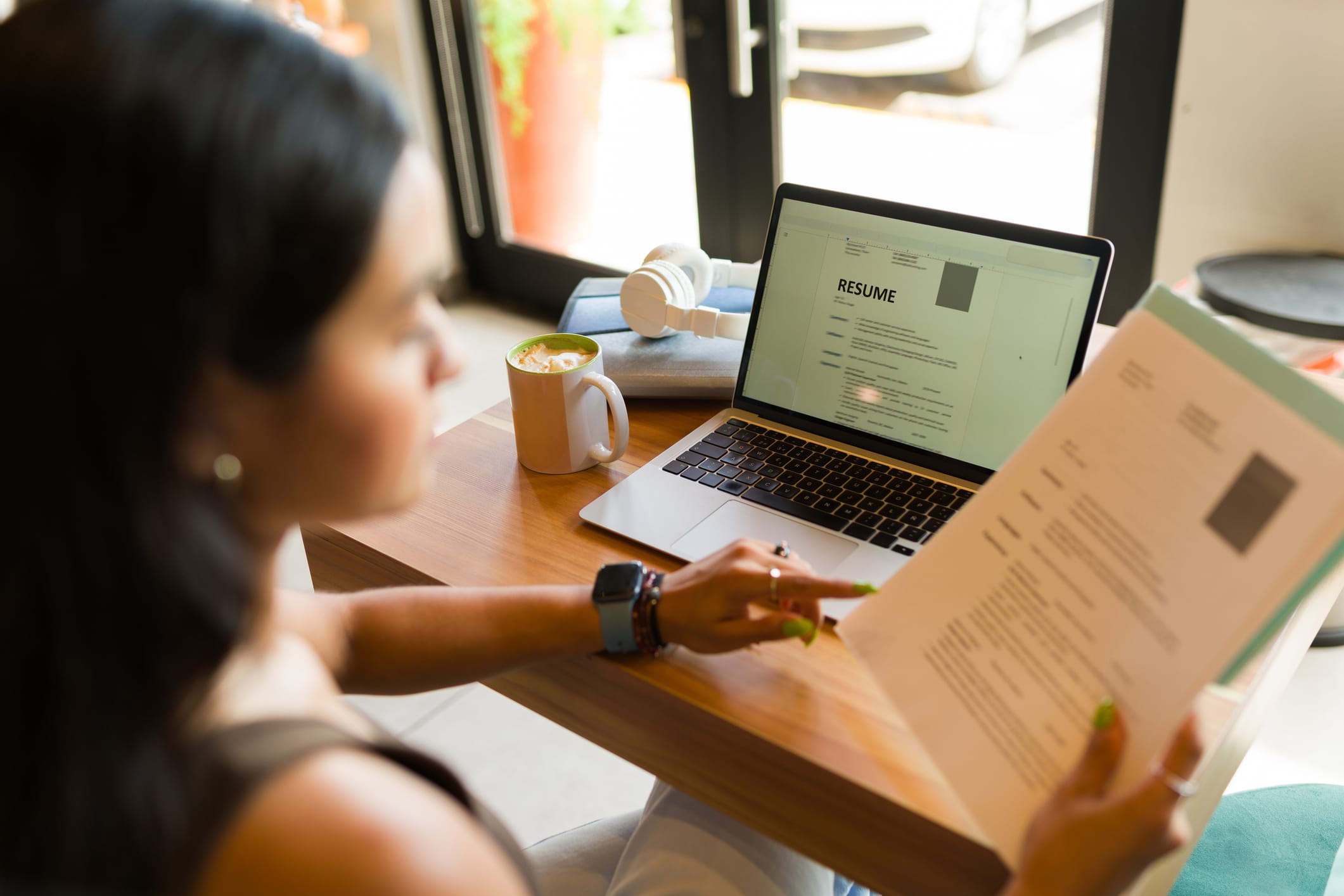 A person reviews a printed resume while sitting at a wooden table, with a laptop and a cup of beverage in a cozy café setting.